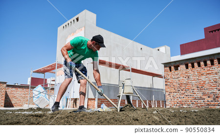 Full length of male worker shoveling sand-cement mix while working at construction site. Man in shorts preparing floor screed material while standing outdoors near concrete screed mixer machine. Full length of male worker shoveling sand-cement mix while working at construction site. Man in shorts preparing floor screed material while standing outdoors near concrete screed mixer machine. 90550834