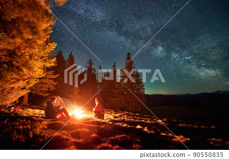 Pair of tourists spending evening time near bonfire on the middle of glade under open fullstar sky with Milky way in the mountains. Tent, trees, mountain peaks on the background. Pair of tourists spending evening time near bonfire on the middle of glade under open fullstar sky with Milky way in the mountains. Tent, trees, mountain peaks on the background. 90550835