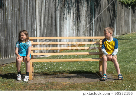 Children boy and girl are in social distancing sitting on bench in park. Brother and sister talking. Kids separated by social distancing. Sad child have to be separated because of the sick 90552196