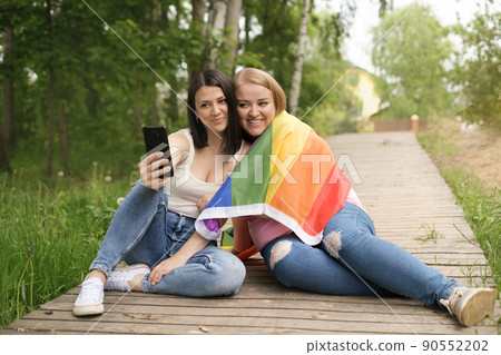 Two lesbian friends are photographed together in a park with an LGBT flag on their shoulder. June is the Month of pride and equality of human rights. Happy cute girls with a phone in hand Two lesbian friends are photographed together in a park with an LGBT flag on their shoulder. June is the Month of pride and equality of human rights. Happy cute girls with a phone in hand 90552202