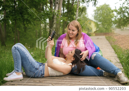 A young brunette woman lies on her girlfriend's lap and shows a video on her smartphone. Two lesbian girls are relaxing outdoors. Rainbow LGBT Flag on girlfriend's shoulder 90552203