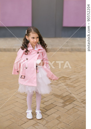 A cute curly-haired girl in an elegant festive dress and a pink raincoat with a broken cast arm looks with a smile towards copy space. A cute curly-haired girl in an elegant festive dress and a pink raincoat with a broken cast arm looks with a smile towards copy space. 90552204