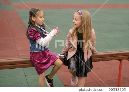 A girl with a cast on her arm is trying to put lipstick on her lips. The child laughs at his helpless girlfriend. Funny kids are sitting on a playground in the city. Friendship of two sisters A girl with a cast on her arm is trying to put lipstick on her lips. The child laughs at his helpless girlfriend. Funny kids are sitting on a playground in the city. Friendship of two sisters 90552205