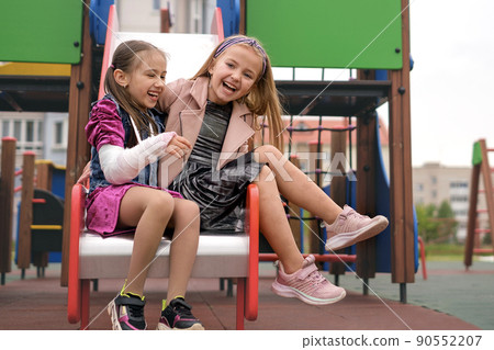 Two funny amusing cool sisters laugh sincerely and have fun on the playground. The girl supports her friend with a hand injury and a cast. Children have fun together on summer vacations Two funny amusing cool sisters laugh sincerely and have fun on the playground. The girl supports her friend with a hand injury and a cast. Children have fun together on summer vacations 90552207
