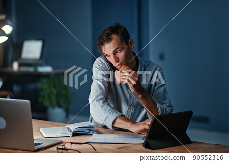 young businessman snacking on a hamburger while working on a new project. 90552316