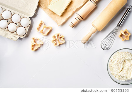 Bowl with wheat flour, rolling pin, whisk, eggs, butter, cookie cutters. Top view on a white table with a copy space 90555393
