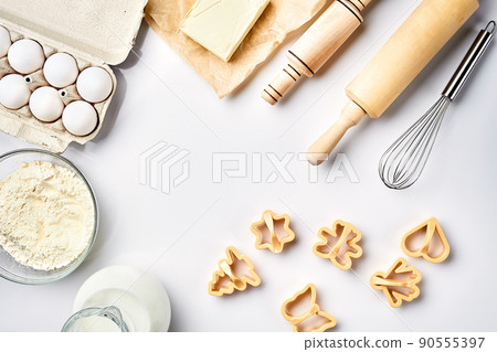 Objects and ingredients for baking, plastic molds for cookies on a white background. Flour, eggs, rolling pin, whisk, milk, butter, cream. Top view, space for text Objects and ingredients for baking, plastic molds for cookies on a white background. Flour, eggs, rolling pin, whisk, milk, butter, cream. Top view, space for text 90555397