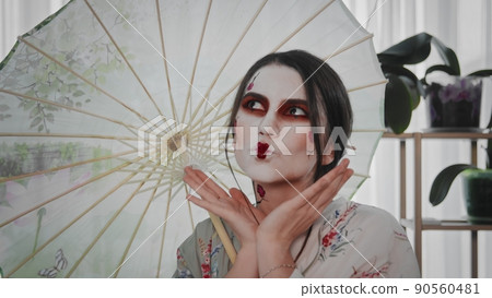 A young beautiful Japanese-style girl stands with an umbrella and a fan in her hands and smiles cheerfully. Portrait of a stylish beautiful geisha under an umbrella with a sincere smile. Image of a A young beautiful Japanese-style girl stands with an umbrella and a fan in her hands and smiles cheerfully. Portrait of a stylish beautiful geisha under an umbrella with a sincere smile. Image of a 90560481