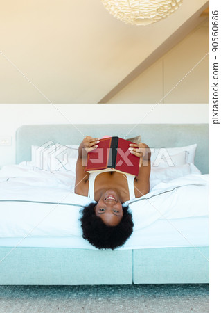 Portrait of african american young woman smiling while holding a book lying on the bed at home 90560686