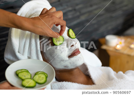 Hands putting cucumber slices over eyes of african american woman with facial beauty mask at home 90560697