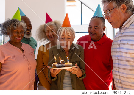 Happy multiracial friends wearing party hats looking at senior woman blowing birthday candles 90561262