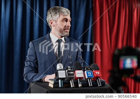 Mature bearded businessman standing at stage with microphones and giving an interview during online broadcast at press conference Mature bearded businessman standing at stage with microphones and giving an interview during online broadcast at press conference 90566540