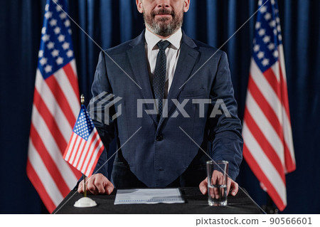 Portrait of male politician giving a speech standing on a stage with American flag behind his back Portrait of male politician giving a speech standing on a stage with American flag behind his back 90566601