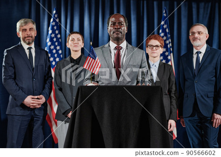 Portrait of African mature leader standing at tribune at press conference with his business team behind him 90566602