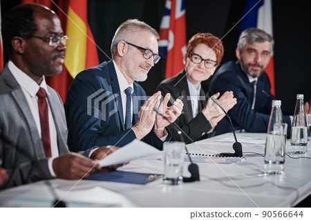 Group of people applauding speech of their colleague while sitting at table during conference 90566644