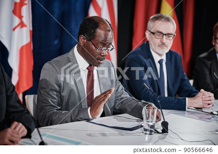 African politician in eyeglasses sitting at table with microphone among his colleagues and reading report during press conference 90566646