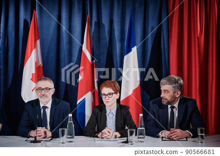 Group of politicians sitting at table during press conference and answering media questions with flags in background Group of politicians sitting at table during press conference and answering media questions with flags in background 90566681