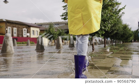 girls in raincoats and rubber boots walk along road flooded with torrential rains, their feet walk through puddles city, splashing water to the sides, the flood is on street, car is driving on water. girls in raincoats and rubber boots walk along road flooded with torrential rains, their feet walk through puddles city, splashing water to the sides, the flood is on street, car is driving on water. 90568790