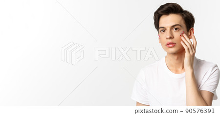 People, lgbtq community and lifestyle concept. Beautiful young gay man applying glitter under eyes for pride party, standing over white background 90576391