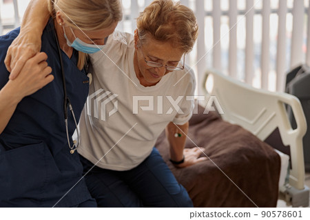 Female doctor in mask helping patient to get up from bed in the ward at hospital Female doctor in mask helping patient to get up from bed in the ward at hospital 90578601