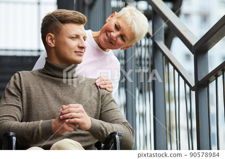 Positive kind female caregiver touching shoulder of patient in wheelchair and talking to him outdoors 90578984