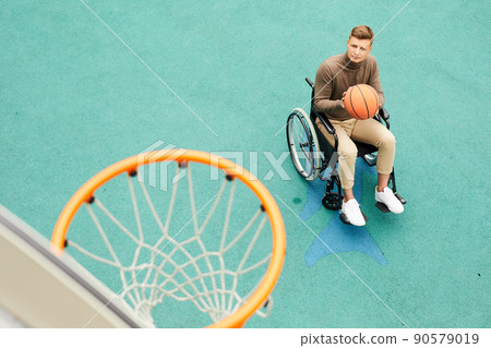 Serious young handicapped man sitting in wheelchair and playing basketball on sports ground Serious young handicapped man sitting in wheelchair and playing basketball on sports ground 90579019