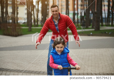 Cheerful father enjoys his day off, spends time with his kid- adorable little girl riding a push scooter in a city park. Happy family relationships concept 90579421