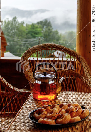 Hot fruit tea in a glass teapot and cookies stands on the edge of a rattan table. Hot fruit tea in a glass teapot and cookies stands on the edge of a rattan table. 90579732