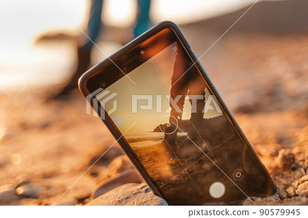 The concept of the photos on the phone. The smartphone lies buried in the sand on the beach, and takes a photo of a man in the background. Close up The concept of the photos on the phone. The smartphone lies buried in the sand on the beach, and takes a photo of a man in the background. Close up 90579945