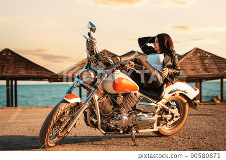 Cool confident adult woman in leather jacket and boots posing lying on motorcycle. Beach umbrellas on the background. The concept of Motorcyclist Day and feminism 90580871