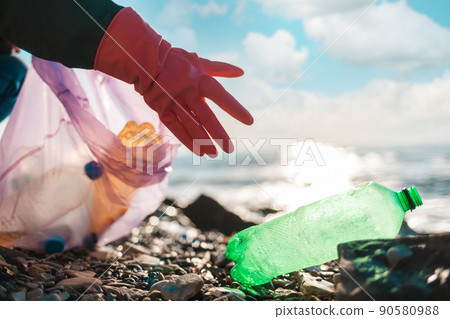 Earth Day. A person in protective rubber gloves reaches for a dirty plastic green bottle lying on the ocean shore. Hand close-up. Copy space. Concept of save the Planet 90580988