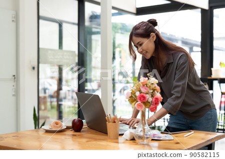 Cheerful attractive young female graphic designer smiling and working on laptop computer at her desk in modern office Cheerful attractive young female graphic designer smiling and working on laptop computer at her desk in modern office 90582115