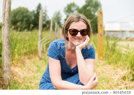 Portrait of an attractive thirty year old girl with a blue dress and sunglasses sitting at the Flemish countryside Portrait of an attractive thirty year old girl with a blue dress and sunglasses sitting at the Flemish countryside 90585038