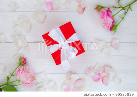 On a white wooden background there are roses and petals forming a circle, inside there is a gift in a red box and a white ribbon. High quality photo On a white wooden background there are roses and petals forming a circle, inside there is a gift in a red box and a white ribbon. High quality photo 90585859