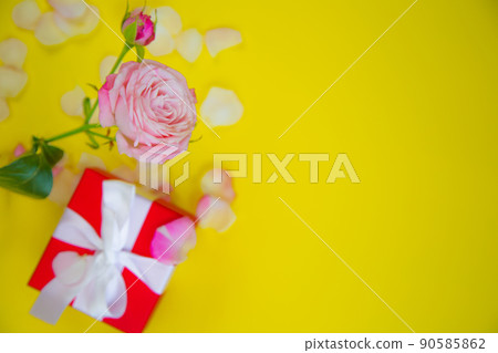 A rose flower and a gift in a red package with a white ribbon stands on the side on a yellow background surrounded by petals with a place for text. High quality photo A rose flower and a gift in a red package with a white ribbon stands on the side on a yellow background surrounded by petals with a place for text. High quality photo 90585862