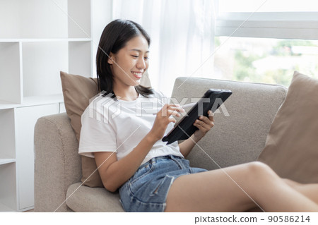 Asian woman sitting on the sofa in the living room using a tablet, Looking at tablet screen, Relaxing at home, Working through the Internet communication system, Happy working lifestyle. 90586214