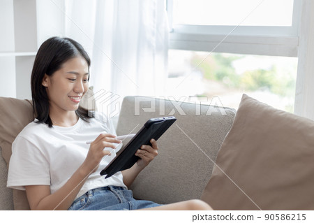 Asian woman sitting on the sofa in the living room using a tablet, Looking at tablet screen, Relaxing at home, Working through the Internet communication system, Happy working lifestyle. Asian woman sitting on the sofa in the living room using a tablet, Looking at tablet screen, Relaxing at home, Working through the Internet communication system, Happy working lifestyle. 90586215