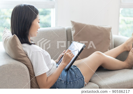 Asian woman sitting on the sofa in the living room using a tablet, Looking at tablet screen, Relaxing at home, Working through the Internet communication system, Happy working lifestyle. 90586217