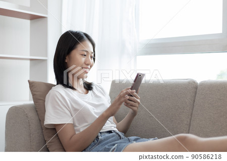 Asian woman sitting with smart phone on sofa in living room, Looking at tablet screen, Relaxing at home, Working through the Internet communication system, Happy working lifestyle. 90586218