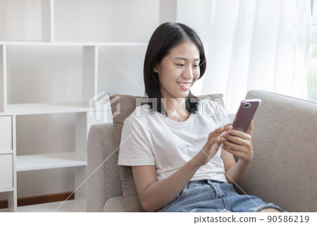 Asian woman sitting with smart phone on sofa in living room, Looking at tablet screen, Relaxing at home, Working through the Internet communication system, Happy working lifestyle. 90586219