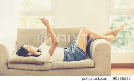 Asian woman sitting with smart phone on sofa in living room, Looking at tablet screen, Relaxing at home, Working through the Internet communication system, Happy working lifestyle. 90586222