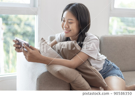 Asian woman sitting with smart phone on sofa in living room, Looking at tablet screen, Relaxing at home, Working through the Internet communication system, Happy working lifestyle. Asian woman sitting with smart phone on sofa in living room, Looking at tablet screen, Relaxing at home, Working through the Internet communication system, Happy working lifestyle. 90586350