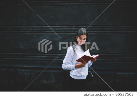 Positive young lady turning page of book while standing near old wall 90587502