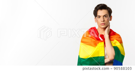 Waist-up shot of beautiful gay man with glitter on face, wrap himself with rainbow lgbt flag and touching face gently, looking at camera, white background Waist-up shot of beautiful gay man with glitter on face, wrap himself with rainbow lgbt flag and touching face gently, looking at camera, white background 90588103
