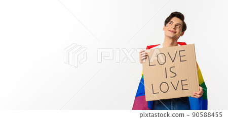 Dreamy young queer person smiling and looking at upper left corner, holding love is love sign for pride parade, wearing Rainbow flag, white background 90588455