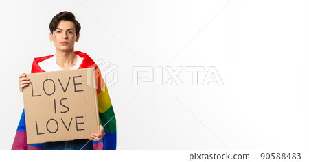 Serious and confident gay man wearing rainbow lgbt flag, holding sign for pride parade, standing over white background 90588483