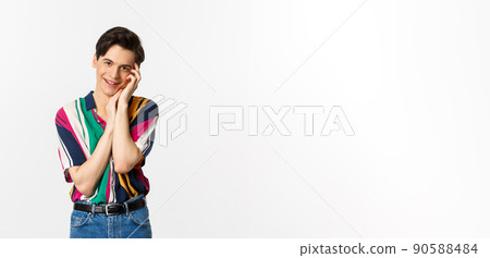 Beautiful young man looking with admiration at camera, smiling, gently touching face, standing over white background 90588484