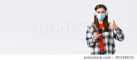 Social distancing, lifestyle on covid-19 outbreak, coronavirus concept. Cheerful pleased good-looking woman in medical mask, approve nice sound of wireless headphones, thumb-up, hold phone 90588656