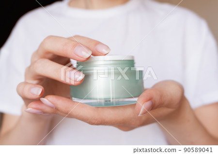 Female hands with natural pink manicure holding a transparent cream jar. Woman in white holds beauty product. Concept of body care and skin protection. Moisturizing lotion closeup. Facial skincare 90589041