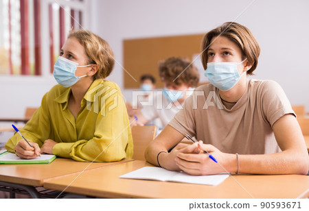 Teenager in protective mask studying in classroom, listening to lecturer and writing in notebook Teenager in protective mask studying in classroom, listening to lecturer and writing in notebook 90593671